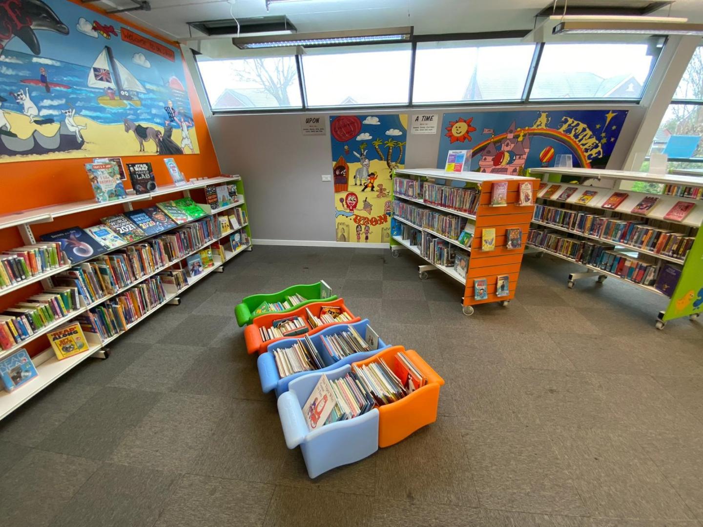 A side view of the children's area at a library, with orange and green shelves and book bins